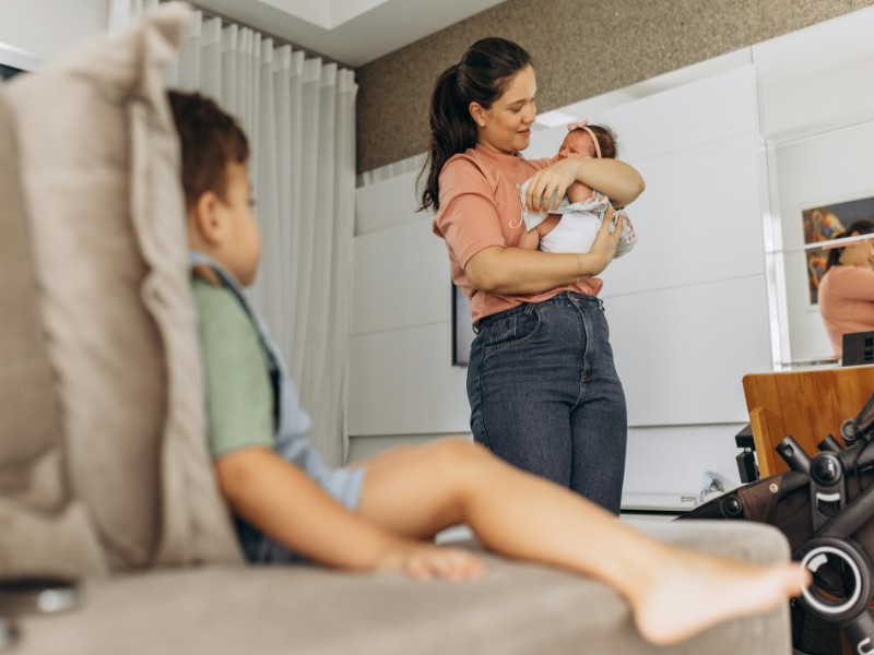 A Filipino childcare service holds newborn while a toddler sits on the couch in a cozy living room setting