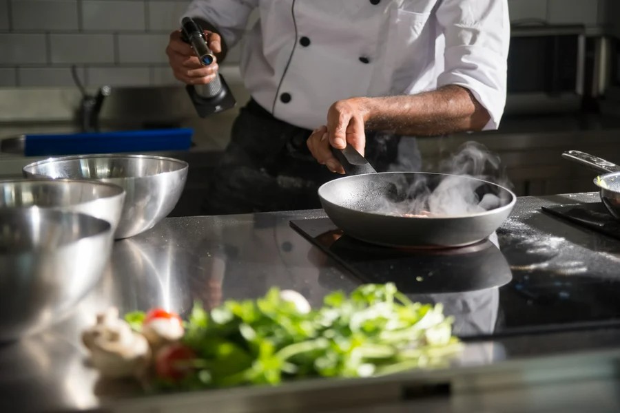 professional cook service prepares food in a sleek kitchen using a frying pan on an induction cooktop